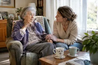 An elderly woman and her adult daughter talk calmly in a bright, cozy living room, holding hands, with a caring and reassuring atmosphere.